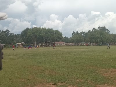 This image depicts a wide-angle view of a large, open grassy field under a heavily clouded sky in Chevaywa ward, Kenya. The foreground consists of uneven green grass, with a partial view of a hand or finger visible in the upper left corner of the frame.
In the midground, a group of approximately 10-12 individuals, appearing to be young adults or children, are gathered on the left side of the field. Several wear red shirts, while others are in blue and yellow attire. One individual in a red shirt is in motion, moving towards the left. Further to the right in the midground, a solitary individual in a red shirt is positioned centrally, facing right. Another individual in a light-colored shirt and dark pants stands towards the far right, with arms slightly raised.
The background features a dense horizontal line of green trees, approximately 8-10 meters tall, separating the field from various single-story buildings. Visible structures include buildings with distinct red-tiled roofs and other lighter-colored buildings, suggesting a mix of residential or institutional architecture. Utility poles and overhead lines span horizontally across the background, situated above the tree line and structures. The overall scene suggests an outdoor gathering or activity, possibly a sports game, on the large field.