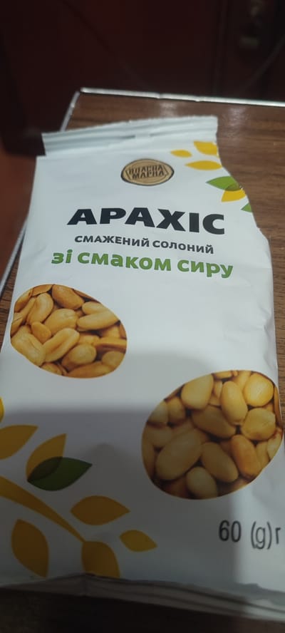 The image shows a bag of Arachis peanuts with a cheese flavor. The bag is white with black and green text, and features images of peanuts and stylized leaves. The text on the bag is in Ukrainian. The bottom right corner of the bag displays "60 (g) r", indicating the weight of the product. The bag appears to be resting on a wooden surface. The location is not discernible from the image.