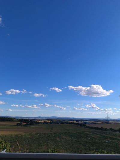 The image depicts a scenic panoramic view of a rural landscape under a clear blue sky with scattered white clouds. The foreground shows a grey metal barrier, likely part of a road or viewpoint, with some greenery visible below it. The middle ground features rolling hills covered in a patchwork of agricultural fields, with varying shades of green and brown indicating different crops or stages of growth. Trees are sparsely distributed across the fields, and a few buildings and industrial silos can be observed in the distance. A prominent pylon with power lines stands to the right of the frame, suggesting the presence of electrical infrastructure. The background consists of layers of hazy blue hills extending to the horizon. The overall impression is one of a serene countryside setting, possibly on the outskirts of Předotice, Czechia, given the agricultural nature of the terrain and the presence of typical European rural elements. No people or specific actions are discernible in the image.