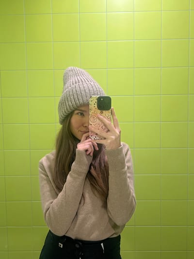 A female subject is centered in a mirror selfie, positioned against a wall covered in square, light green ceramic tiles with visible grout lines. She wears a light grey knitted beanie, a light brown turtleneck sweater with ribbed cuffs, and dark-colored lower garments. Her long, brown hair extends over her shoulders. Her right hand holds a smartphone with a light-colored, celestial-themed case, obscuring the left side of her face and functioning as the camera. Her visible right eye is open and directed forward. Her left hand is raised towards her face, with her index finger touching her lower lip area. A thin ring is present on her left ring finger. The location within Nazarovo, Russia, cannot be ascertained from the visual evidence.