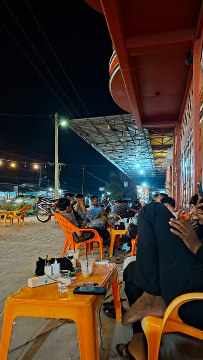 This media file depicts a nighttime scene at an outdoor establishment, likely a cafe or food stall, in Keudeteupinpunti, Indonesia. Several individuals are seated at bright orange plastic chairs, some facing the camera and others looking away. The subjects are diverse in age and attire, with some dressed casually and others in more formal wear. Their body language suggests a relaxed atmosphere, with some engaged in conversation, while others are engrossed in their phones or simply observing their surroundings.
The setting is characterized by an overhead awning supported by a metal structure, creating a covered area for patrons. The ground appears to be paved with interlocking bricks. Various objects are visible, including plastic cups, mobile phones, lighters, and power strips on tables, suggesting a place where people gather to eat, drink, and socialize. A motorcycle is parked nearby, hinting at transportation methods.
The time of day is clearly nighttime, with artificial lighting casting a warm glow on the scene. The weather appears to be clear, as there are no indications of rain or extreme temperatures. The overall mood is one of casual enjoyment and social interaction. Although no prominent text is visible, the presence of multiple people in a communal space strongly implies a public gathering or a casual dining experience.