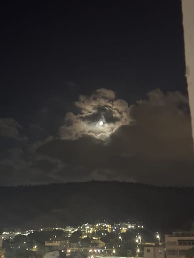 The image captures a nighttime view of Tiberias, Israel, with the full moon partially obscured by clouds. The moon is the prominent celestial object, casting a soft glow through breaks in the atmospheric haze. Below the sky, a dark silhouette of a hill or mountain range dominates the midground, its contours indistinct against the night sky. The foreground reveals a spread of city lights, indicating a residential or urban area. Buildings are visible, characterized by illuminated windows and streetlights, suggesting a bustling townscape despite the darkness. The overall scene conveys a sense of quietude and the ethereal beauty of a moonlit night in a city.