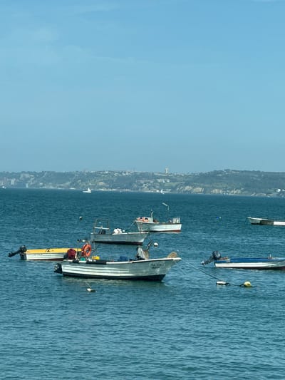 The image depicts a waterfront scene in Oeiras, Portugal, with several boats moored in the blue sea. The boats are of various sizes and colors, with some appearing to be fishing vessels and others smaller recreational boats. The water is calm with slight ripples. In the background, a hilly coastline is visible, dotted with buildings and trees, suggesting an urban or suburban area. The sky is a clear, pale blue with some faint clouds. The overall impression is one of a peaceful, sunny day at sea. The boats are the primary focus, with their reflections shimmering on the water's surface.