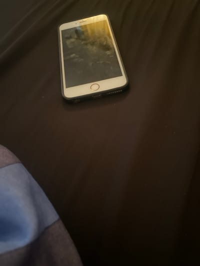 A white/silver smartphone, fitted with a black protective case, rests diagonally on a dark brown or black, slightly textured fabric surface. The phone's screen is dark but exhibits reflections of an overhead light source. The bottom edge of the device, featuring a charging port and speaker grilles, is visible. In the lower left quadrant of the image, a partial segment of blue and dark striped fabric is present. The specific location in the city of Katcha, Nigeria, or any other geographical detail, cannot be determined from the visual information provided. No individuals are depicted, and no explicit actions or interactions are occurring.