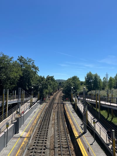 An eye-level, wide-angle shot captures a railway station with parallel tracks running into the distance under a clear blue sky. The station features two platforms with raised walkways connecting them over the tracks. The walkways are lined with metal railings and yellow support poles, with some pedestrians visible on them. Yellow safety lines mark the edges of the platforms. The tracks themselves are surrounded by green trees and vegetation, with overhead power lines and a distant pylon visible. The scene appears to be located in Oviedo, Spain, likely at a functional but not overly busy railway stop.