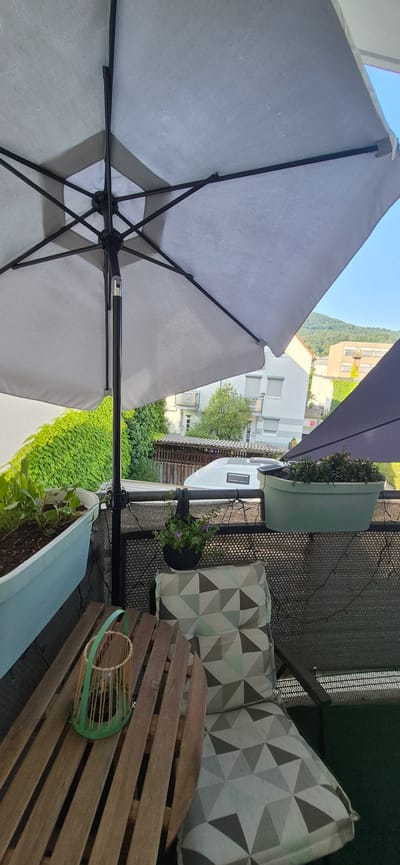 The image depicts a balcony in Baden-Baden, Germany, featuring outdoor furniture and plants. In the foreground, a wooden table with a slatted surface holds a green basket-like candle holder. To the right, a chair is partially visible with a cushion adorned with a geometric pattern of grey and light blue triangles.
Behind the table and chair, a balcony railing is covered with a dark mesh screen. Hanging from the railing is a black pot containing small purple flowers. To the left, a long, light blue planter box holds green plants.
Overhead, a large, light gray umbrella is open, with its dark pole and metal frame visible. Partially obscured behind the umbrella, a dark navy blue umbrella is also present.
In the background, a dense green hedge lines the edge of the property. Beyond the hedge, buildings with light-colored facades and shuttered windows are visible, along with trees. A white recreational vehicle is parked in the middle ground, with a fence visible in front of it. Hills covered with trees form the distant background, under a clear blue sky. The overall impression is a well-maintained outdoor space, likely a residential balcony, in a European setting.
