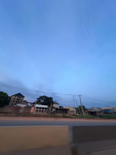 The image captures a roadside view in Orifite, Nigeria, under a vast, pale blue sky with wispy clouds. The foreground is dominated by a greyish-brown barrier, suggesting a road or elevated pathway. Beyond this barrier, a paved road with a darker asphalt surface stretches horizontally across the frame. Along the far side of the road, a collection of buildings and structures are visible.
On the left, a multi-story building under construction, with a dark, sloped roof and visible window frames, stands prominently. Adjacent to it are lower buildings, some appearing to be shops or homes with corrugated metal roofs. A large tree with dense green foliage is situated behind these structures. Further to the right, more buildings with various roof designs are visible, some appearing to be simple, single-story structures. Utility poles with visible power lines are spaced along the roadside, indicating infrastructure. The environment appears to be somewhat rural or suburban, with a mix of ongoing construction and established dwellings. The lighting suggests it may be either early morning or late evening. No people are clearly discernible in the image.