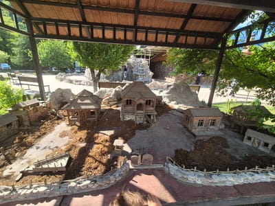 This media file shows a miniature village diorama displayed outdoors under a shaded canopy. The diorama features several small wooden buildings, resembling traditional houses and structures, situated on a landscape of dirt and dried leaves. Miniature fences made of stones and sticks border parts of the scene. In the background, a man-made waterfall cascades over artificial rocks, adding a touch of natural scenery to the display. Beyond the diorama, glimpses of a real-world setting are visible, including trees, parked cars, and paved pathways, suggesting the diorama is part of a larger park or attraction. The lighting indicates it is daytime, likely a sunny day given the strong shadows. There is no discernible activity or event taking place within the diorama itself, other than its static display. Visible text on the underside of the canopy appears to be branding or manufacturer information, but it is mostly illegible and fragmented.