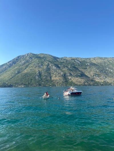 This image captures a daytime scene in the Bay of Kotor, near Prčanj, Montenegro, characterized by calm teal-blue waters and imposing mountains under a clear sky.
The upper two-thirds of the frame consists of a cloudless, vibrant blue sky. Below, a large, verdant mountain range dominates the background, stretching across the horizon. The mountains are covered with a mix of dense green foliage and exposed grey rock formations.
The foreground and midground depict a calm body of water, displaying a gradient of deep teal and blue, indicating depth and clarity. Two boats are central to the scene. On the left, a small, white rowing boat contains two individuals. One person, wearing a red garment, is seated and engaged in rowing. Another person is seated in the bow.
Further to the right and slightly behind the rowing boat, a larger white motorboat with a dark hull stripe and red fenders is positioned. At least three individuals are visible on the deck of this motorboat, some standing. One person is in the water near the stern of the motorboat, with their head visible above the surface. Fishing rods extend from the stern. The name "ADRIANA" and a partial registration number "PR" are visible on the boat's side.
Separately, in the lower-left foreground, another individual's head is visible, partially submerged in the water. A yellow buoy is partially visible in the far-right midground. The overall impression is one of leisure activities on a sunny day in a picturesque, mountainous bay.
