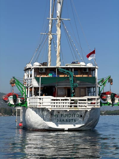 The image shows the stern of a ship named "MV PINDITO JAKARTA" docked in Buleleng, Indonesia. The ship is a working vessel, possibly a cargo or research ship, with multiple cranes, rigging, and sails visible. A red and white Indonesian flag is flying from a mast. The ship is in calm water, reflecting the blue sky and surrounding greenery. The time of day appears to be late morning or early afternoon, with bright sunlight casting shadows. The overall impression is one of a bustling port or a peaceful day at sea.

The ship itself is white with a grey hull. It has a multi-level deck with various equipment and cargo containers. Two people are visible on the deck, one operating a crane and the other appears to be inspecting equipment. The water is a deep blue and shows ripples from the boat's movement. In the distance, lush green hills can be seen, suggesting a tropical location.

The name of the ship, "MV PINDITO JAKARTA," is clearly visible on the hull. The Indonesian flag signifies the country where the ship is located or registered. The presence of cranes and rigging suggests that the ship is actively involved in some form of maritime operation, whether it's loading and unloading cargo, scientific research, or fishing. The overall atmosphere is serene and tranquil, with the gentle movement of the water and the calm demeanor of the people onboard.