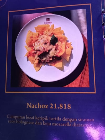 This image captures a section of a menu, predominantly dark blue with a gold border. The central subject is a photograph of a plate of "Nachoz," served on a white, round plate. The nachos consist of tortilla chips generously covered with a reddish bolognese sauce and melted, bubbly mozzarella cheese. A sprig of fresh green herb, possibly parsley or cilantro, garnishes the dish. The word "cenato" is visible in red text on the rim of the plate, indicating a brand or restaurant name. The plate is set on what appears to be a warm-toned wooden surface.

Below the image, in a golden-brown font, the dish is labeled "Nachoz 21.818," which includes its price. Further text in Indonesian, "Campuran lezat keripik tortila dengan siraman saos bolognese dan keju mozarella diatasnya," describes the dish. This translates to "Delicious mixture of tortilla chips with a drizzle of bolognese sauce and mozzarella cheese on top." The image is a typical menu display, aiming to showcase the food appealingly. The presence of Indonesian text and the given location context suggest this menu is from a establishment in Situbondo, Indonesia. The right edge of the menu page shows punched holes, implying it is part of a bound menu book.