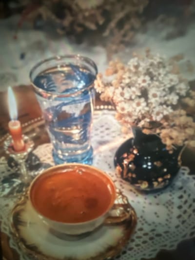 A vintage still life features a clear, etched blue glass lying horizontally next to a saucer and teacup filled with orange liquid. A small black pitcher with gold accents sits nearby. Delicate white flowers are arranged in a vase behind the glass, all resting on a white lace tablecloth. A small, lit candle in a holder is visible at the bottom. The image has a slightly out-of-focus, dreamlike quality.