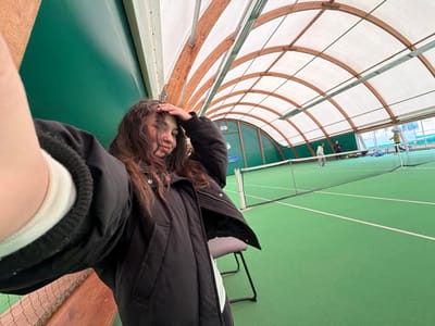 This image depicts an indoor tennis court facility located in Souffelweyersheim, France. In the immediate foreground, a young female is captured in a selfie-style photograph. She has long, wavy dark brown hair and is wearing a black puffer jacket over a dark undergarment, with a white cuff visible on her left sleeve. Her left hand is raised to her head/hair, and she displays a subtle smirk with her left eye appearing partially closed. A dark chair with a white element is partially visible in the lower left foreground.
The background showcases a green indoor tennis court with white boundary lines. A white tennis net spans the width of the court. The facility's ceiling is an arched structure supported by wooden beams, covered with translucent white panels that allow for natural light. Additional fluorescent lighting fixtures are suspended from the ceiling. The side walls of the facility are green.
Beyond the net, three individuals are present on the court. One person, in light-colored attire, stands near the far baseline. A second person, dressed in darker clothing, is positioned further back on the court to the right. A third person, wearing a yellow top, is visible towards the right side of the far court. Several blue objects, possibly equipment bags, are situated along the back wall of the court area.