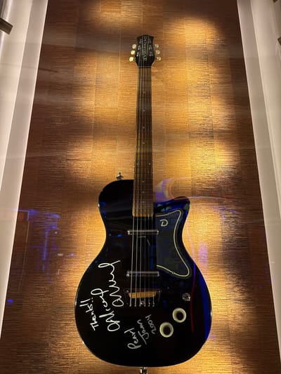 The image depicts a black electric guitar mounted on a brown textured wall. The guitar has signatures in white ink, including "Pearl Jam 2009" and "Thanks!! Alice in Chains." The guitar is under a glass display case, and the location is in Hollywood Seminole Reservation, United States of America.