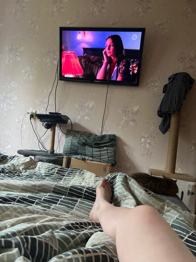 The image depicts an indoor scene, likely a bedroom, with a woman's legs and feet prominently in the foreground, suggesting she is lying on a bed. The bed is covered with a duvet featuring a geometric pattern of grey and white squares. In the background, a flat-screen television is mounted on a wall covered with floral wallpaper. The TV displays a scene from a movie or show, featuring a woman. To the left of the TV, a cat tree is visible, with electrical wires and a power outlet connected to devices on its platforms. A striped shirt is draped over a post of the cat tree. A tabby cat is resting on a box behind the bed, partially obscured. The presence of floral wallpaper and the style of the furniture suggest a domestic setting in a residential area. The specific location within the city of Tula, Russia, cannot be determined from the visual information alone.