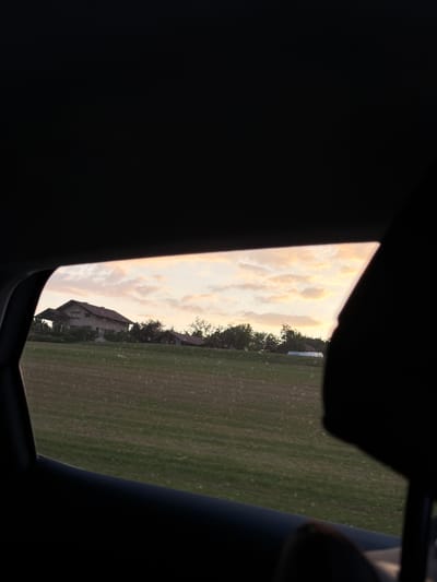 The image captures a view from inside a vehicle, looking out a window. The foreground is dominated by the dark interior of the car, including a silhouette of a headrest in the right portion of the frame. Beyond the window, a field of green grass stretches out under a sky filled with soft, pale clouds illuminated by a setting sun. In the distance, a house with a dark roof and light-colored walls is visible, surrounded by trees. The location is identified as being in the city of Groisy, France. The overall scene suggests a peaceful, rural landscape during twilight.