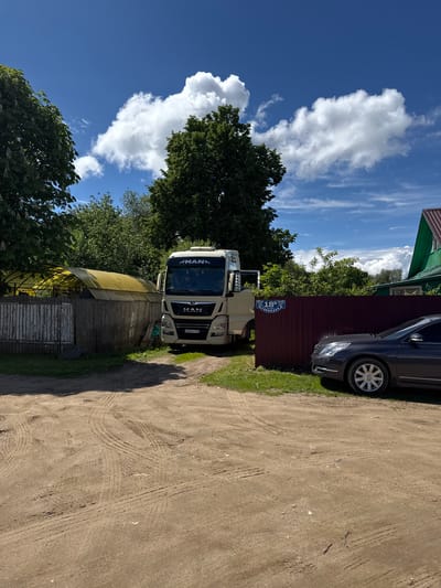 The image depicts a dirt road in what appears to be a rural or semi-rural area of Slobodka, Russia. A white MAN truck is parked alongside a fence with an address plaque reading "18 Б". A dark-colored sedan is parked further along the fence. A shed-like structure is visible near the truck. The sky is blue with scattered clouds, and trees are visible in the background.