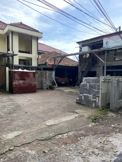 A narrow alleyway sits between buildings. Piles of bricks, some brown and some gray, are stacked along the edges. Overhead, a tangle of electrical wires cuts across a cloudy sky. Buildings with tiled roofs line the alley, showing a mixture of architectural styles and varying levels of upkeep. The pavement is cracked and uneven, with gravel along the edges.