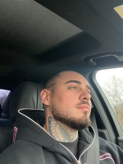An adult male is depicted in a medium close-up, seated in the front passenger or driver's seat of a vehicle in Mikre, Bulgaria. The individual has light skin, a short buzz cut, dark, defined eyebrows, and a neatly trimmed full beard and mustache. A stylized feather or leaf tattoo is visible on his left temple, and extensive black ink, including what appears to be a large butterfly or moth design, covers his neck. He is wearing a black zippered hoodie with pink accents and a black seatbelt across his right shoulder and chest. His gaze is directed upwards and to the right, looking out the vehicle's side window. The car's interior features a dark, textured headliner, a visible sunroof panel, and black seating. Through the window, an exterior view of natural foliage and trees is visible, suggesting a rural or forested area under overcast lighting conditions.