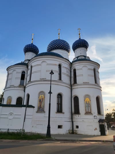 The image shows a white church with blue domes decorated with gold stars, against a blue sky with some clouds. The church has multiple levels and arched windows. There are golden mosaics of religious figures on the exterior walls. A black lamppost stands in front of the church. A paved walkway and a street are visible in the foreground, with some grass and a low fence along the edge of the walkway. In the background, trees and a few distant figures and vehicles are discernible. This structure is identified as the Church of the Transfiguration in Chelganovo, Russia.