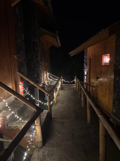 The image shows a night scene in Du Già Commune, Vietnam. A narrow stone path, illuminated by strings of small white lights, leads between wooden buildings. The structures have stone and wood construction, with visible roofs and support beams. The sky is dark, suggesting night. The scene is still and without visible people or movement.