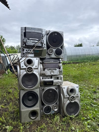In Elovo, Russia, a tall, haphazard stack of discarded or stored silver-colored mini hi-fi stereo components and speakers is positioned outdoors on a grassy, uneven patch of ground under an overcast sky.

The stack comprises multiple units from different manufacturers. At the top left, an LG-branded DVD Karaoke system, model number partially obscured, features a blank rectangular screen, a large central volume knob, and an array of smaller buttons and ports. Below it, a unit with a cassette deck slot is visible, partially open, with internal components exposed. To its right, a silver LG speaker has a large main driver, a smaller mid-range driver, and a tweeter.

Further down and to the right, a sequence of stacked main stereo units includes one with a "3 CD CHANGER" label and another with "MDSS" branding, each presenting various control buttons, small display screens, and input/output interfaces. Wires, predominantly black, are loosely draped across and through these central units.

At the mid-left, a Samsung-branded three-way speaker stands, featuring a large circular woofer at the bottom, a smaller mid-range driver, and a bass reflex port above it. A similar Samsung speaker, oriented with the woofer at the top and the port at the bottom, is positioned at the lower left. Another identical Samsung speaker is located at the lower right. All speakers feature protective grilles over their drivers. The metal casing of these components exhibits dust accumulation and minor surface wear.

The immediate ground surrounding the stack is covered with sparse green grass, patches of exposed dirt, and various weeds, including dandelions and broad-leafed plants. In the background to the left, a light-colored wooden handle, possibly from a shovel, is visible leaning near a dark-colored wheelbarrow. In the middle distance to the right, a large, translucent, semi-cylindrical greenhouse or polytunnel spans horizontally. Beyond the greenhouse, cultivated fields or garden plots are discernable, interspersed with green vegetation. The distant horizon features a line of green deciduous trees under a uniformly grey, cloudy sky. No human presence or dynamic action is depicted.