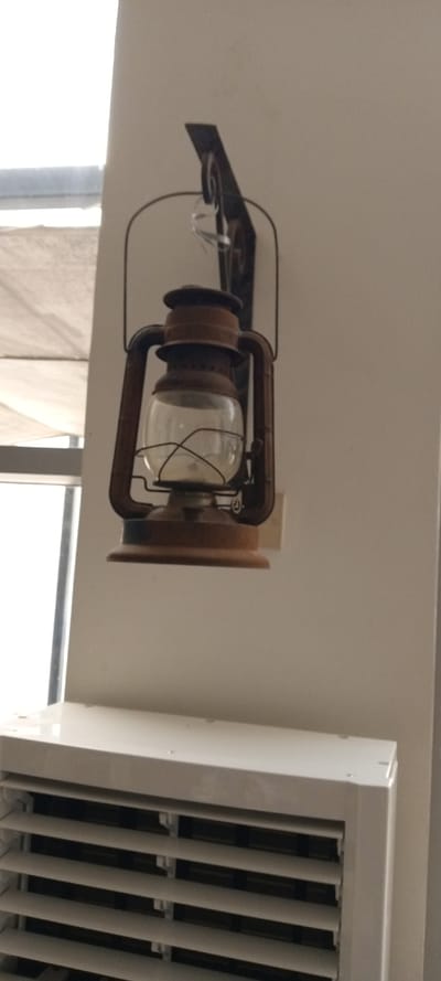 An image displays an indoor scene in Yola, Nigeria, focused on a rustic lantern mounted on a light-colored wall. A brown, oil-lamp style lantern with a clear glass globe is suspended from a dark, possibly wrought-iron, decorative wall bracket. The bracket is affixed to an off-white or beige painted wall. To the right of the lantern's base, a light-colored electrical outlet is visible on the wall. Below the lantern, the upper portion of a white, louvered air conditioning unit or cooler is present. To the far left, a window frame allows diffused daylight into the room. There are no individuals present, and no discernable actions or interactions are occurring.