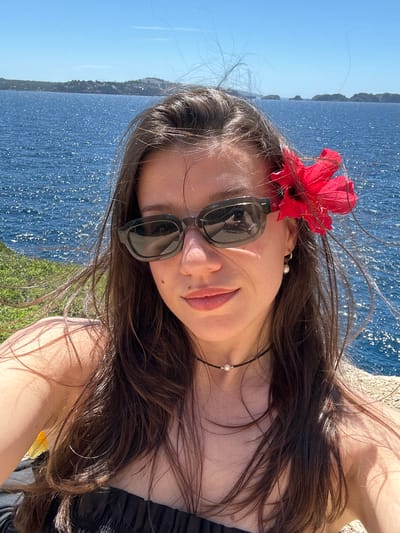 A young woman is posing for a selfie on what appears to be a grassy bank or edge, with a large body of water visible in the background. The water is sparkling under a clear blue sky. On the opposite shore, trees are visible. The woman has long, dark hair, and a red flower is clipped to the side of her head. She is wearing sunglasses and a delicate necklace with a pearl. Her facial expression is relaxed and she is angled towards the camera.
Without additional information or metadata associated with the image, pinpointing a specific location in Calvià, Spain, is not possible. Calvià has an extensive coastline with many similar viewpoints.