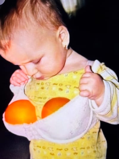 The image shows a close-up of a baby wearing a yellow, patterned onesie. The baby has light-colored hair and is looking downwards. The baby's hands are near its mouth and chest. A white bra is positioned in front of the baby's torso, containing two round, orange objects. The background is dark and out of focus. There are no discernible landmarks or features that would indicate a specific location in Beșalma, Moldova.