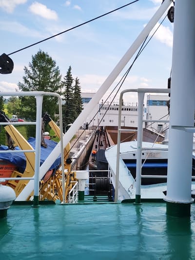 The image shows a view from the deck of a ship, looking towards a canal lock. The deck is painted a glossy teal color and reflects the sky. In the foreground, there are white railings and parts of the ship's structure. A yellow crane-like apparatus is visible on the left, covered in a blue tarp. In the middle ground, a lock gate is visible, with water on both sides. Another ship, largely white with a brown cargo area, is positioned within the lock. A white building with blue window frames stands on the right side of the lock. Tall green coniferous trees are seen on the left bank of the canal, with more distant buildings and a red car visible behind them. The sky is blue with scattered white clouds. The scene appears to be set in a non-urban, possibly industrial or port area. The city of Белоусово, Russia is mentioned in the prompt, however, the image itself does not contain any explicit visual cues to confirm or deny this specific location.