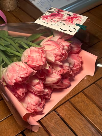 A bouquet of approximately ten to twelve pink double-petal tulips rests on a slatted wooden surface, possibly a table, indoors in Slavyansk-na-Kubani, Russia. The tulips are arranged with their green stems and leaves visible beneath the flower heads. The bouquet is wrapped in a layer of clear plastic film over light pink paper or foil. A small rectangular card with pink floral illustrations and partially visible Cyrillic text, including "МАРТА" and the numeral "8," is clipped to the clear plastic wrapping using a small green plastic clip. In the blurred background, a spool of natural-colored twine is visible to the left, and a notebook or papers with handwritten text are visible to the right. The scene is illuminated by an overhead light source, casting subtle shadows. No individuals are present or engaged in any direct action.