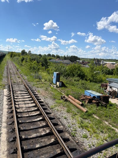 This is a wide shot of a railway track stretching into the distance, bordered by overgrown vegetation and scattered industrial debris. The sky above is a vibrant blue, dotted with fluffy white clouds, suggesting a clear and pleasant day. The scene appears to be set in an industrial or semi-abandoned area, possibly on the outskirts of a town, given the distant buildings and structures visible beyond the trees.
The railway tracks themselves are made of metal rails supported by concrete sleepers, many of which are stained and weathered. Adjacent to the tracks, there is a patch of rough terrain covered in gravel and grass, with several large, rusty pipes lying on the ground. Further back, amidst the dense green foliage, there are glimpses of industrial equipment and possibly some corrugated metal structures. In the background, more buildings and what look like piles of gravel or construction material can be seen.
The lighting indicates it is daytime, likely late morning or early afternoon, due to the bright sunlight casting shadows. There are no people visible in the frame, and the overall atmosphere is one of quietness and slight desolation, typical of an industrial site that is no longer in active use or is undergoing a slow transition. The orientation of the photo, being taken from a slightly elevated perspective looking down along the tracks, provides a sense of depth and perspective. The location is identified as Нижняя Мактама, Russia. There is no visible text in the image.