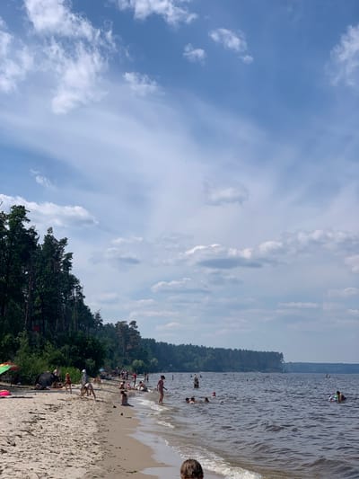 This is a landscape-oriented photograph taken at a beach on a sunny day. The scene depicts a shoreline with a body of water, a sandy beach, and a treeline in the background. A group of people are visible in and around the water, as well as on the beach.

The subjects include numerous individuals, some of whom are swimming or wading in the water, while others are relaxing or playing on the sand. Objects such as a colorful umbrella and a person's head in the foreground are also present. The setting is a natural environment with a clear sky filled with scattered clouds, indicating fair weather. The time of day appears to be daytime, likely afternoon, given the brightness and the shadows.

The activity taking place is recreational, with people enjoying a day at the beach. The overall mood conveyed is one of leisure and enjoyment. The treeline suggests a forested area bordering the beach, and the water appears to be a lake or a calm sea. Location cues like the specific treeline and the nature of the beach could further identify it as Liutizh, Ukraine. There is no visible text in the image.