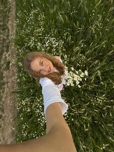 An overhead shot captures a young woman lying in a field of grass and daisies. Her head is tilted back and she smiles at the camera. She is wearing a white top with long sleeves. Her right arm is extended forward, holding a bouquet of daisies. The field is lush and green, with a path on the left side. The location is in the village of Snovitsy, Russia.