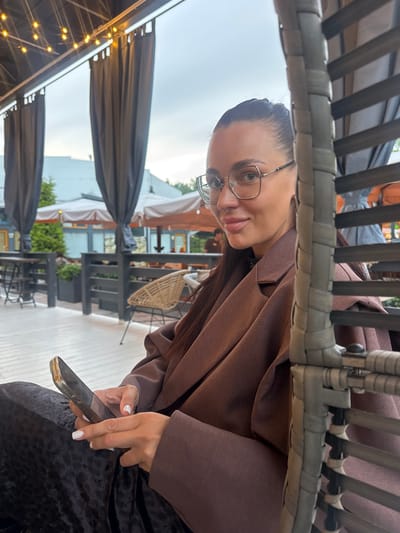 In Vidnoye, Russia, a woman with glasses and a brown coat is seated at an outdoor cafe. She is looking towards the camera with a slight smile. She is holding a dark-colored phone in her right hand. The background shows tables and chairs of the cafe, with umbrellas providing shade. String lights are visible above. The overall atmosphere appears casual and relaxed, possibly during the daytime or early evening. The woman's attire suggests cooler weather.