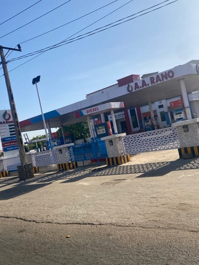 This image depicts the A.A. RANO petrol station in Barnawa, Nigeria, captured from a slightly elevated vantage point across an asphalt road. The station features a prominent white canopy with red and blue stripes, bearing the "A.A. RANO" brand name in red lettering. The word "DIESEL" is visible on the canopy's front edge above a fuel pump island. Behind the pumps, a white building with red accents is visible, labeled "A.A. RANO OFFICE."

A tall, multi-paneled sign pole on the left side of the frame displays "A.A. RANO" at the top, followed by a list of services including "PETROL," "DIESEL," "KERO," "LPG," "RANO Lube," "RANO Autocare," and "RANO Gas." A low barrier separates the station forecourt from the road, constructed with white lattice-patterned sections and blue metal gate segments. The posts supporting this barrier have bases marked with black and yellow stripes. Multiple fuel pumps are discernible under the canopy, predominantly blue and white.

The foreground consists of a dark grey paved road surface showing visible cracks and minor debris. Strong, dark shadows from the station's barrier and structure are cast across the road, indicating direct sunlight from a clear sky. A wooden utility pole with overhead power lines is visible on the far left. In the background, beyond the station, green foliage from trees is visible beneath a bright, clear blue sky. Several individuals are present within the petrol station premises, though their specific actions are not discernible due to distance and resolution.