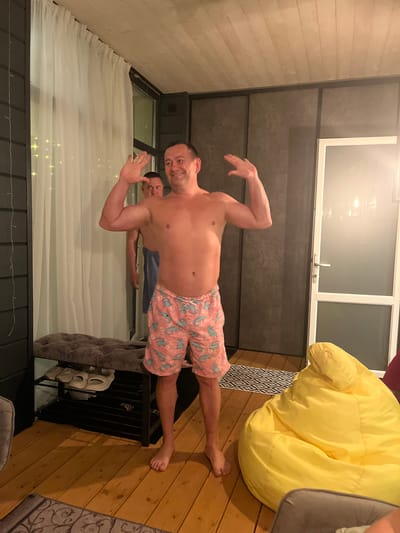 The image shows two men indoors. The man in the foreground is shirtless and wearing pink shorts with a blue dinosaur pattern. He is smiling and has his arms raised. A second man is partially visible behind him, wearing a blue shirt. The setting appears to be a living room, featuring a large window with sheer curtains, a beanbag chair, a rug, and a wooden floor. A shoe rack is partially visible near the center. The location is given as Новая Чернушка, Russia.