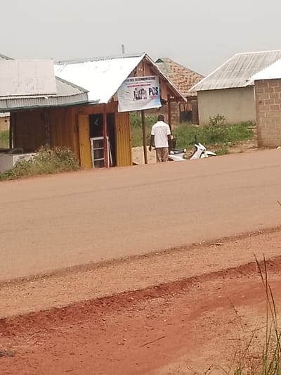 A man in a white shirt and tan pants stands on a dirt road in Agwala, Nigeria. He is facing away from the camera, looking towards a small yellow and brown building with a corrugated metal roof. A white motorcycle is parked beside him. The building appears to be a shop, with a sign that reads "FIONA POS TELECOMMUNICATIONS". Another sign, partially obscured by the man, is also visible. In the background, there are other buildings with corrugated metal roofs, some appearing to be made of brick. The scene is set during the daytime, with overcast skies suggesting a cloudy or possibly rainy weather. The ground is a reddish-brown dirt road, with sparse vegetation on the sides. The overall impression is of a rural or semi-rural commercial area.