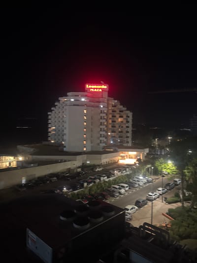 The image shows a nighttime view of the Leonardo Plaza hotel in Tiberias, Israel. The hotel is a large, white building with a distinctive stepped pyramid shape. A red neon sign on top of the hotel reads "Leonardo PLAZA". The hotel is illuminated by lights from its windows and from streetlights. In front of the hotel is a parking lot filled with cars, and there are trees and landscaping around the property. In the background, the lights of Tiberias and the Sea of Galilee are visible. A construction crane can be seen in the upper right portion of the image.