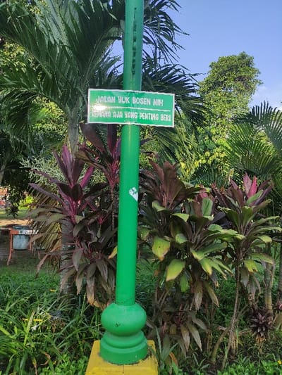 The image shows a green lamppost with a sign attached to it. The sign says "Jalan Yuk Bosen Nih! Laporan Aja Yang Penting Ber2." The lamppost is located in a park setting with lush green grass and various trees. The lamppost is located in Mojokerto, Indonesia, at the coordinates -7.464 latitude, 112.432 longitude. There are no people visible in the image.