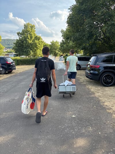 Two young men are standing on a paved surface near parked cars and trees. One man, wearing a light green t-shirt and dark shorts, is pushing a portable cooler with wheels. The cooler is emitting a white smoke or steam. He is positioned to the left of the cooler and is looking away from the camera. Another man, wearing a black t-shirt with an Adidas logo, black shorts, and a black backpack, is walking away from the camera, carrying a white tote bag with red and blue circular designs. He is wearing flip-flops.

Behind them, a black SUV is parked, and another lighter-colored SUV is behind it. Trees and foliage are visible in the background, suggesting an outdoor, possibly park-like setting. The lighting indicates it is daytime, likely sunny with some clouds in the sky. The scene appears casual, possibly a picnic or outdoor gathering. No specific event or emotion is strongly conveyed, other than the general activity of moving the cooler and carrying the bag.

Visible text on the tote bag includes "lohnt sich" and "Lidl". A sign with "Bürgstadt" is partially visible on the car in the background.