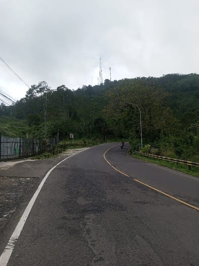 A winding asphalt road with a yellow center line leads uphill towards a lush, green hillside. Trees and vegetation cover the slope, and several tall communication towers are visible near the summit. A guardrail borders the right side of the road. A motorcycle travels along the road. The sky is overcast and gray, casting a soft light over the scene. A metal fence and some greenery are visible on the left side of the road at the bottom of the image.