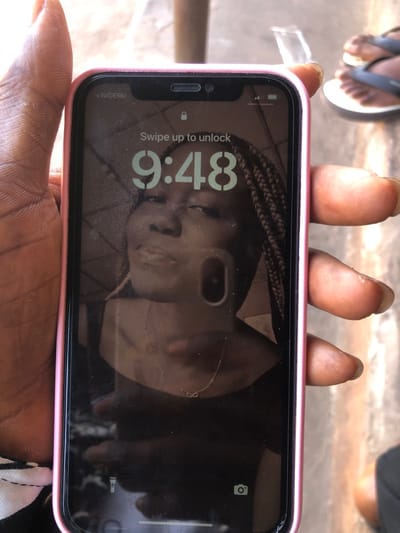 A dark-skinned individual's hand holds a pink-cased iPhone vertically in an outdoor setting in Oraukwu, Nigeria. The phone screen displays the lock screen, showing "NIGERIA" as the carrier, the time "9:48," a padlock icon, and the instruction "Swipe up to unlock." The wallpaper features a dark-haired person with braided hair, wearing a dark top and an infinity symbol necklace. A reflective glare on the screen partially obscures the wallpaper, showing a dark, vertical rectangular object. In the background, to the right of the phone, another individual's feet wearing dark flip-flops with white soles are visible on a light-colored, possibly concrete or tiled, ground. A blurred glass object and a reddish-brown vertical structure are also present in the background.