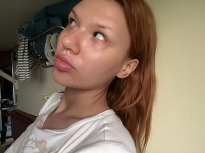 A close-up, eye-level, medium shot shows a young woman with auburn hair and fair skin. She is looking upwards and to her left, with her lips slightly parted. Her head is tilted back, and her right eye is visible, while her left eye is partially obscured. She is wearing a white t-shirt with a light-colored floral print. In the background, a metal rack holding blue towels and white striped fabric is visible, along with a portion of a beige wall. The scene appears to be indoors, in a domestic setting. No specific location within Karmamei, Russia can be determined from the image.