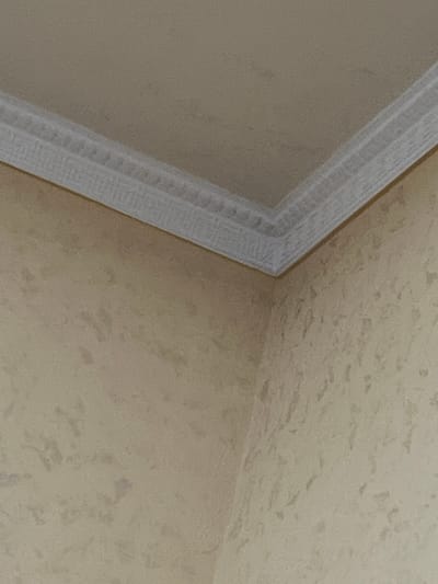 The image displays an interior architectural detail, focusing on a corner where two walls and a ceiling converge. The perspective is upward, showing the junction of these surfaces.
Both the wall (left side) and the ceiling (right side) are painted in a light, warm beige or tan hue. They exhibit a distinct, irregular, and mottled texture, suggesting a decorative plaster or specialized paint finish, with subtle variations in shade creating depth.
A white or off-white crown molding, also known as a cornice, runs diagonally across the frame, marking the seam between the wall and the ceiling. This molding features a series of parallel lines or stepped grooves, indicating a decorative, somewhat ornate design. It appears clean and well-maintained.
There are no people, animals, or specific objects visible in the scene, nor is any activity taking place. The lighting appears soft and uniform, characteristic of an indoor setting. No text is present in the image. The depicted scene is located in Jrarbi, Armenia.