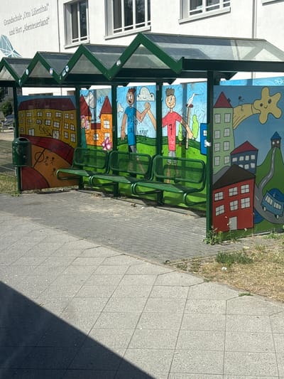 The image shows a covered waiting area with murals painted on the back panels. The murals depict cartoon-like figures and scenes, possibly related to children's activities or stories. One panel shows a yellow giraffe-like creature standing on houses, with a road and a blue bus in the background. Another panel features a person in a red shirt holding a kite and a bird. Below that, there are depictions of houses and people. The bottom panel shows a stylized representation of a playground with stick figures.

The waiting area has green metal benches. A dark green trash can is visible on the left side. The structure is supported by green metal framing.

To the left of the waiting area is a white building with windows. A sign on the building's exterior reads "Grundschule Otto Lilienthal" and "Hort 'Abenteuerland'". This indicates the location is a school and after-school care facility.

The ground in front of the waiting area is paved with grey tiles. To the right, there is a strip of dry grass and weeds. The lighting suggests it is daytime, with sunlight casting shadows on the pavement, indicating a sunny day. The overall atmosphere appears cheerful due to the colorful murals. The image is taken from a slightly angled perspective, with the camera oriented vertically.