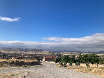 This is a wide-angle, eye-level outdoor shot of a rural landscape on a bright day with a clear blue sky and scattered white clouds. In the foreground, a dirt road curves uphill, flanked by dry, yellow grass. A power line runs horizontally across the top left of the image. In the middle ground, a small town with red-tiled roofs and beige buildings is nestled into the terrain. Beyond the town, a range of dry, rocky hills stretches across the horizon. The overall impression is one of a sunny, tranquil, and sparsely populated area.