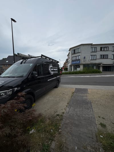 This is a full, eye-level, outdoor, daytime, medium shot of a black Mercedes-Benz Sprinter van parked on a gravel shoulder in front of a street and a light-colored building. The van, which appears to be a work van for an electrical company named "de Ianche," has a ladder rack and a ladder on its roof. The building in the background has multiple windows and the word "COPPER" is visible on a blue sign. A pedestrian walkway made of brick is visible in the foreground. The sky is overcast. The scene is located on the Steenokkerzeelsestraat in Wemmel, Belgium. There are no people visible in the image.