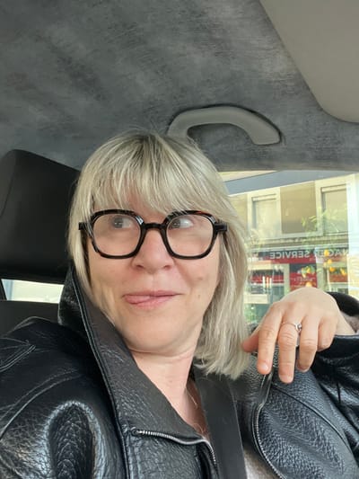 A blonde-haired woman with dark-rimmed, tortoiseshell glasses is seated within a vehicle, partially obscured by a black headrest to her left. Her eyes are directed upwards and to the right, and her tongue is slightly extended from the left corner of her mouth. She wears a black, textured leather jacket with a visible silver zipper along the left lapel. A silver-colored ring is present on the ring finger of her left hand, which rests on the jacket. The vehicle's grey, textured headliner and a grey grab handle are visible above her. Reflected in the vehicle's window to her right is a street view from Veauche, France, specifically depicting the building housing "BANHAGE SERVICE" located at 21 Rue de Saint-Étienne. The mirrored reflection shows "ECIVRES EGAHNAB" on a red and white storefront sign, beneath which a retail display, possibly containing citrus fruits, is also reflected. Above the storefront, multiple building windows are visible in the reflection.