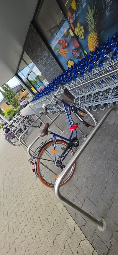 In Freren, Germany, a line of blue shopping carts is parked outside a building with a large glass window displaying an advertisement for fruits and vegetables. In front of the shopping carts, several bicycles are parked in a metal rack. The bicycles are of various colors and styles, with one blue bicycle in the foreground having a basket and a brown seat. The ground is paved with interlocking gray stones. The scene appears to be in front of a store or supermarket.